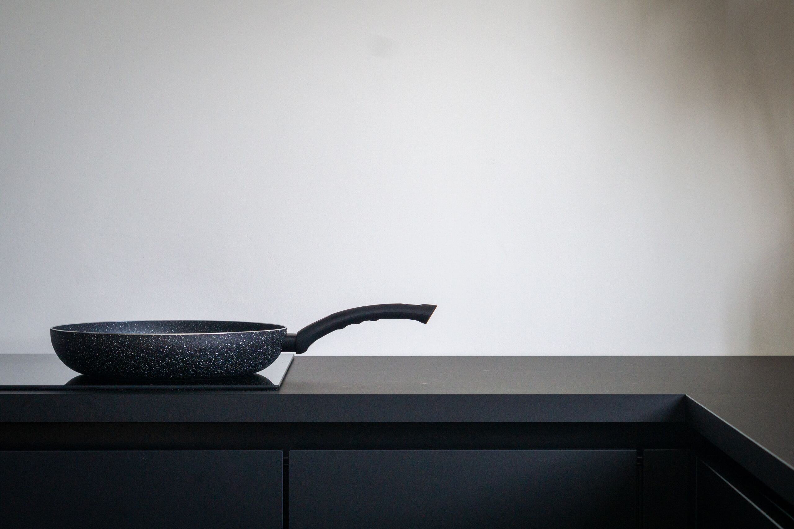 The image shows a black frying pan with a speckled pattern, placed on a black stovetop or countertop. The pan has a long, ergonomic handle. The background is a plain white wall, creating a minimalist and clean aesthetic. The scene is simple and uncluttered, focusing solely on the frying pan, which appears to be new and unused. The overall atmosphere is modern and sleek, with a strong emphasis on the contrasting colors of black and white.